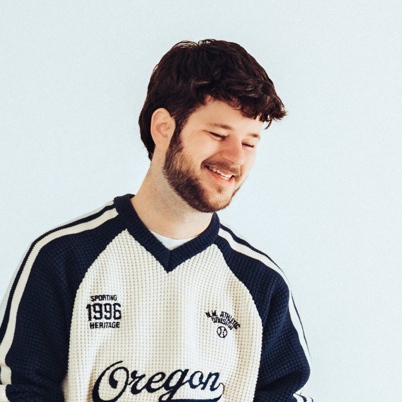 APRD, DJ and producer from Utrecht, smiling in a cream Oregon varsity sweater against a light background