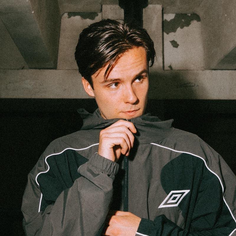 LUMINE, Dutch producer and DJ, in a grey and black Umbro track jacket against a concrete wall