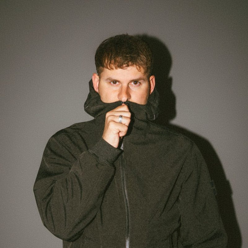 RSCL, Dutch DJ and producer Franklin van Beek, pulling a dark green hoodie up over his mouth against a grey wall